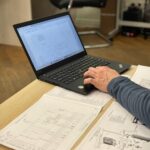 An engineer works on a laptop reviewing technical blueprints and diagrams in an office.